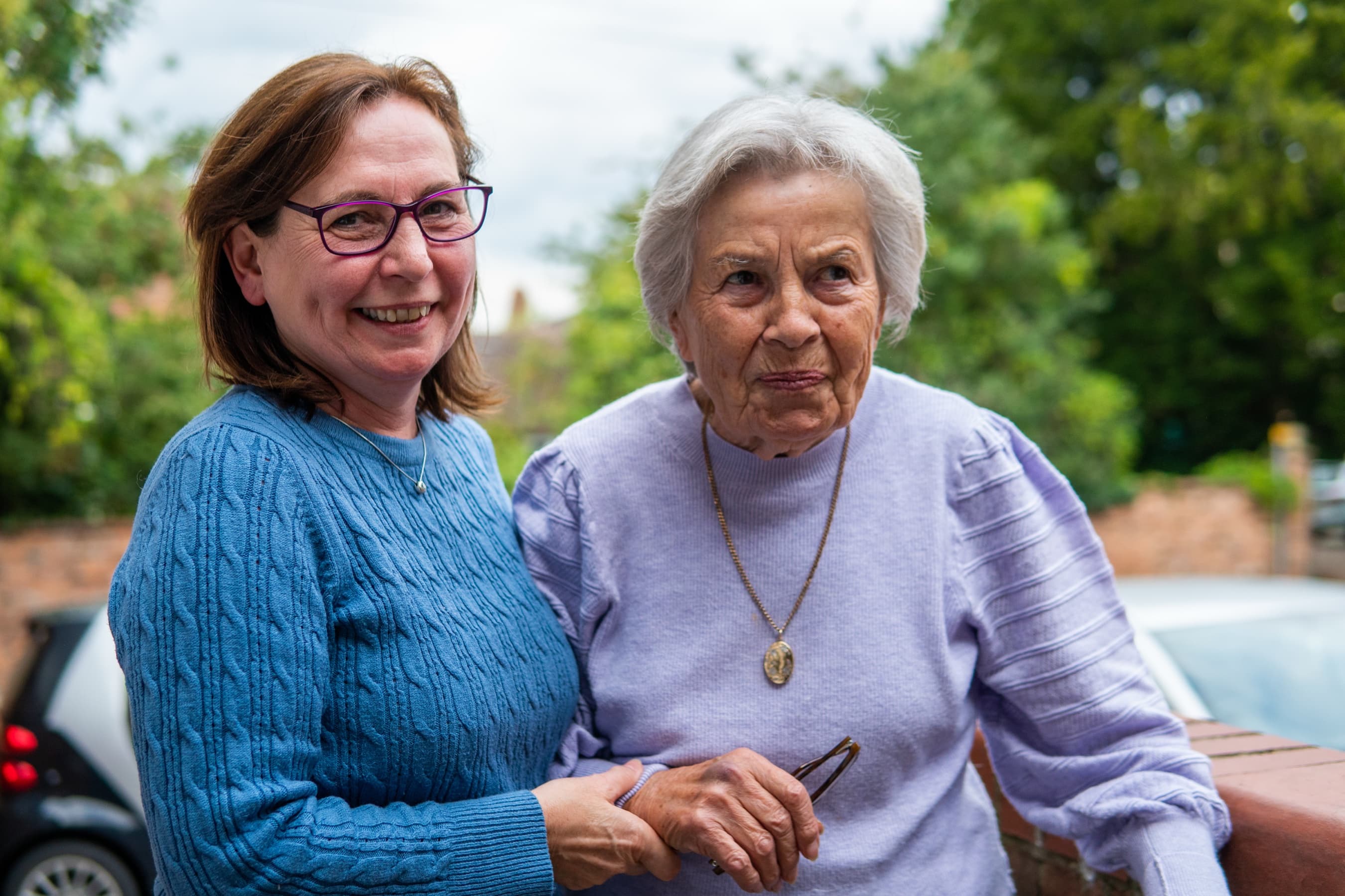 A woman stands with her arm through her mother's, both looking towards the camera.