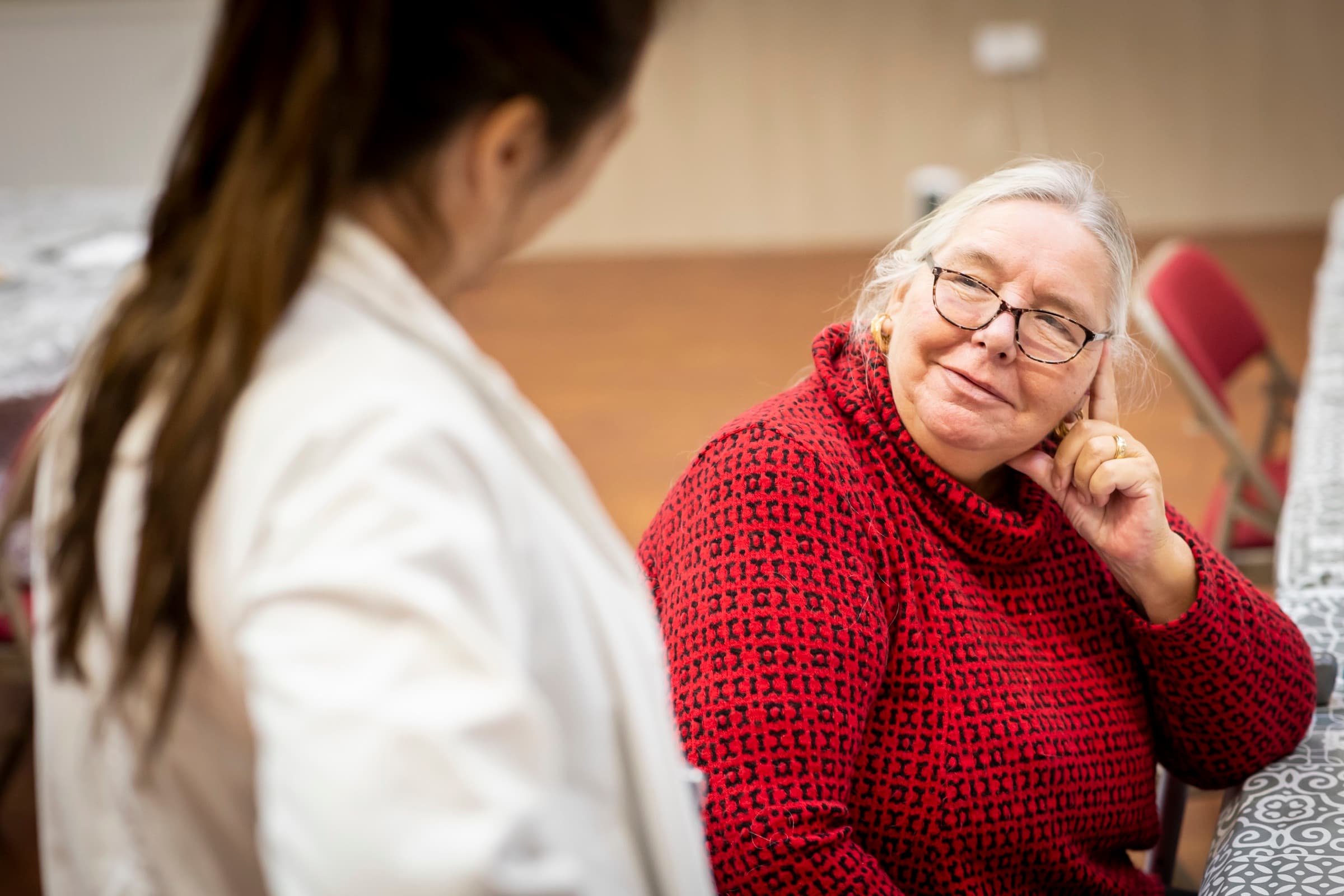 An older woman in a red jumper smiles as she talks with someone.