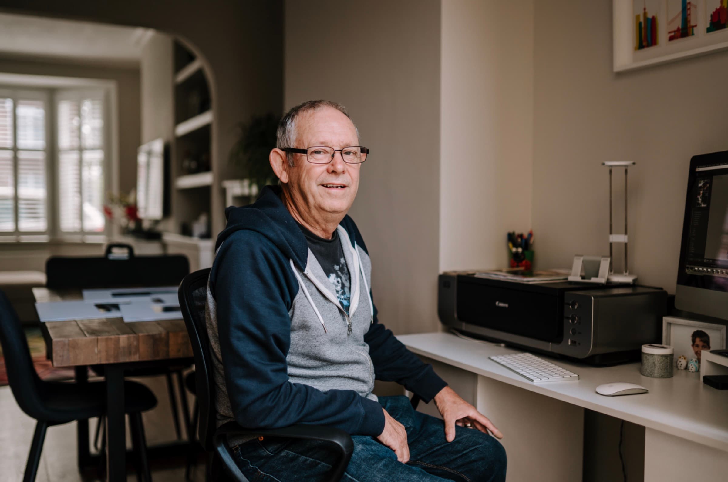 An older man sits at a desk in his home, smiling at the camera.
