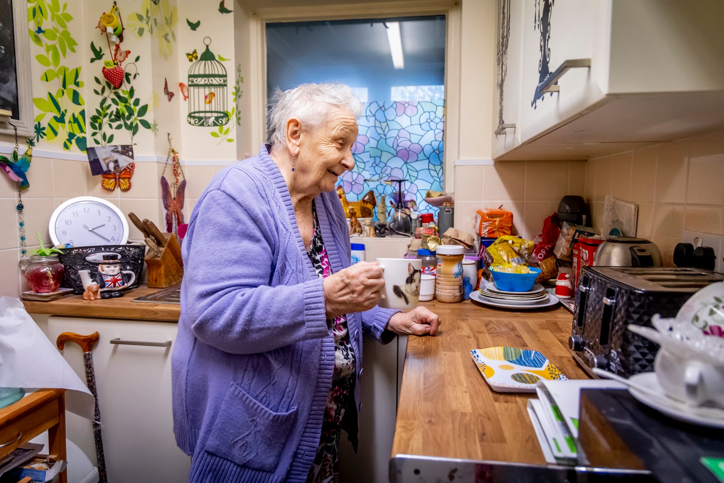An older woman stands in her own kitchen holding a mug, smiling, with her walking stick nearby.